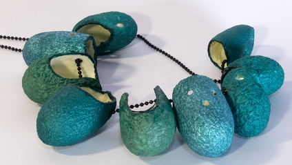 Halskette mit 9 aufgefädelten, vorne offenen Seidenkokons, die außen smaragdgrün, innen mit Nachtleuchtpigment bemalt sind