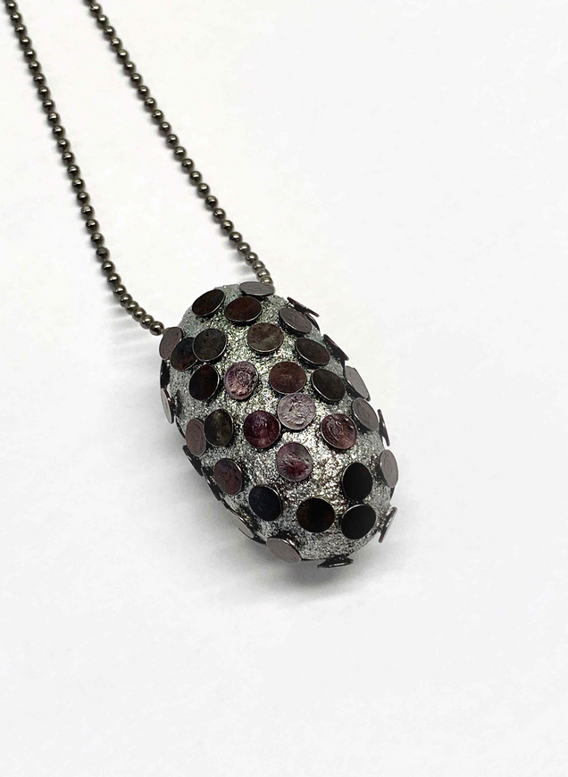 Geschlossener silberner Seidenkokon, in den viele flache schwarze Nägel gestochen sind, an einer schwarzen Silberkette