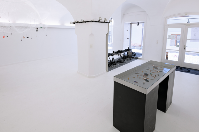 The exhibition Mulberry Tubes by jewellery artist Michaela Rapp shows an installation of havy and small tubes on a low bench on a grey paper that is placed on the wall and the floor; cocoon necklaces inside a see-through net hanging down from the ceiling and a black vitrine with glass.