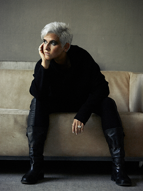 Ela Nord Woman with silverwhite hair, black pullover, black pants and black boots, she looks to the left, her head resting relaxed in the hand